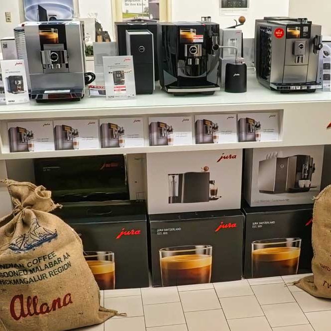 Kaffeevollautomaten Regal mit drei modernen Jura-Kaffeemaschinen, jeweils mit Infokarte davor. Darunter gestapelte Produktkartons mit Jura-Logo und Abbildungen