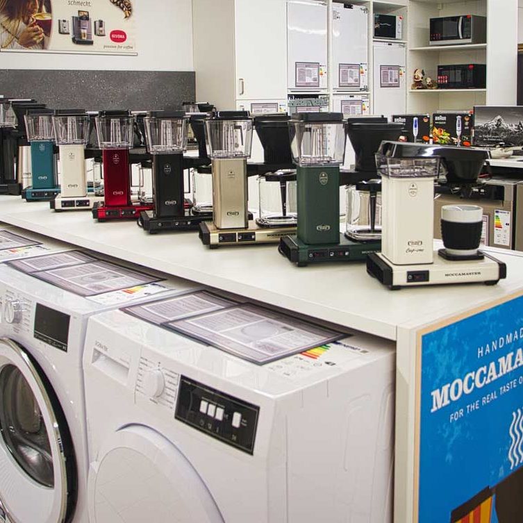 Qualität und Vielfalt Verschiedene Moccamaster Kaffeemaschinen in verschiedenen Farben auf einem Verkaufstisch. Darunter Waschmaschinen. Im Hintergrund Mikrowelle